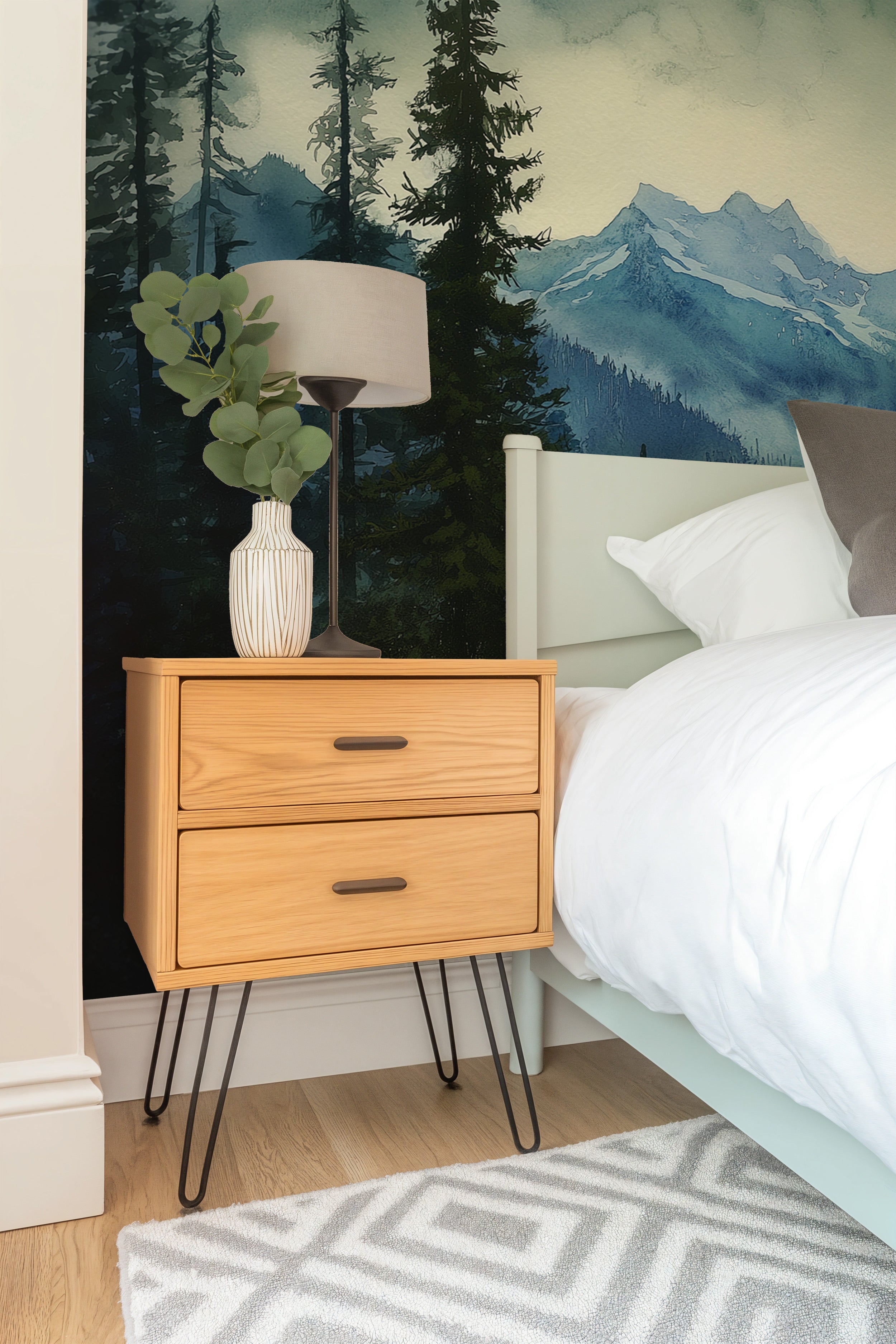 Watercolour mountain forest mural in a bedroom with a wooden two-drawer nightstand, potted greenery, and a light bed against