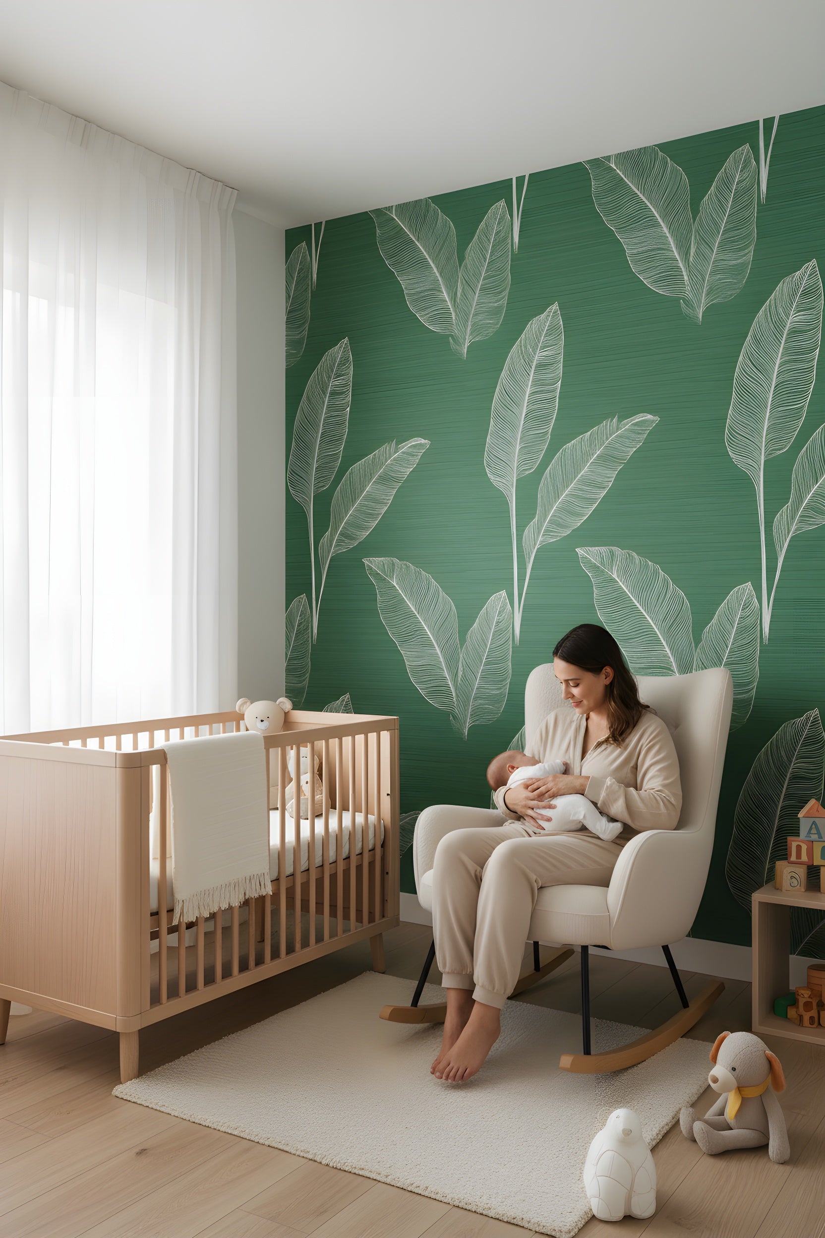 A nursery scene featuring emerald green tropical leaf wallpaper with white outlines behind a wooden crib and a white rocking chair where a mother