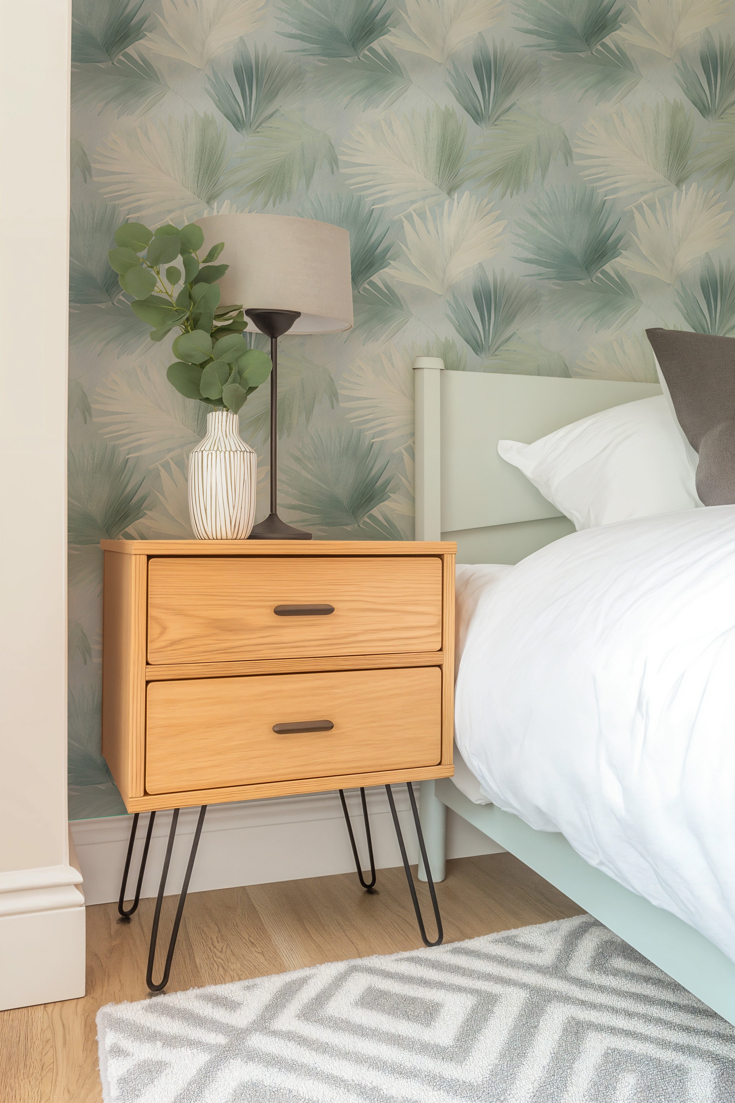 Tropical palm leaves wallpaper in sage green and cream install backdrop beside a light wood two-drawer nightstand with black