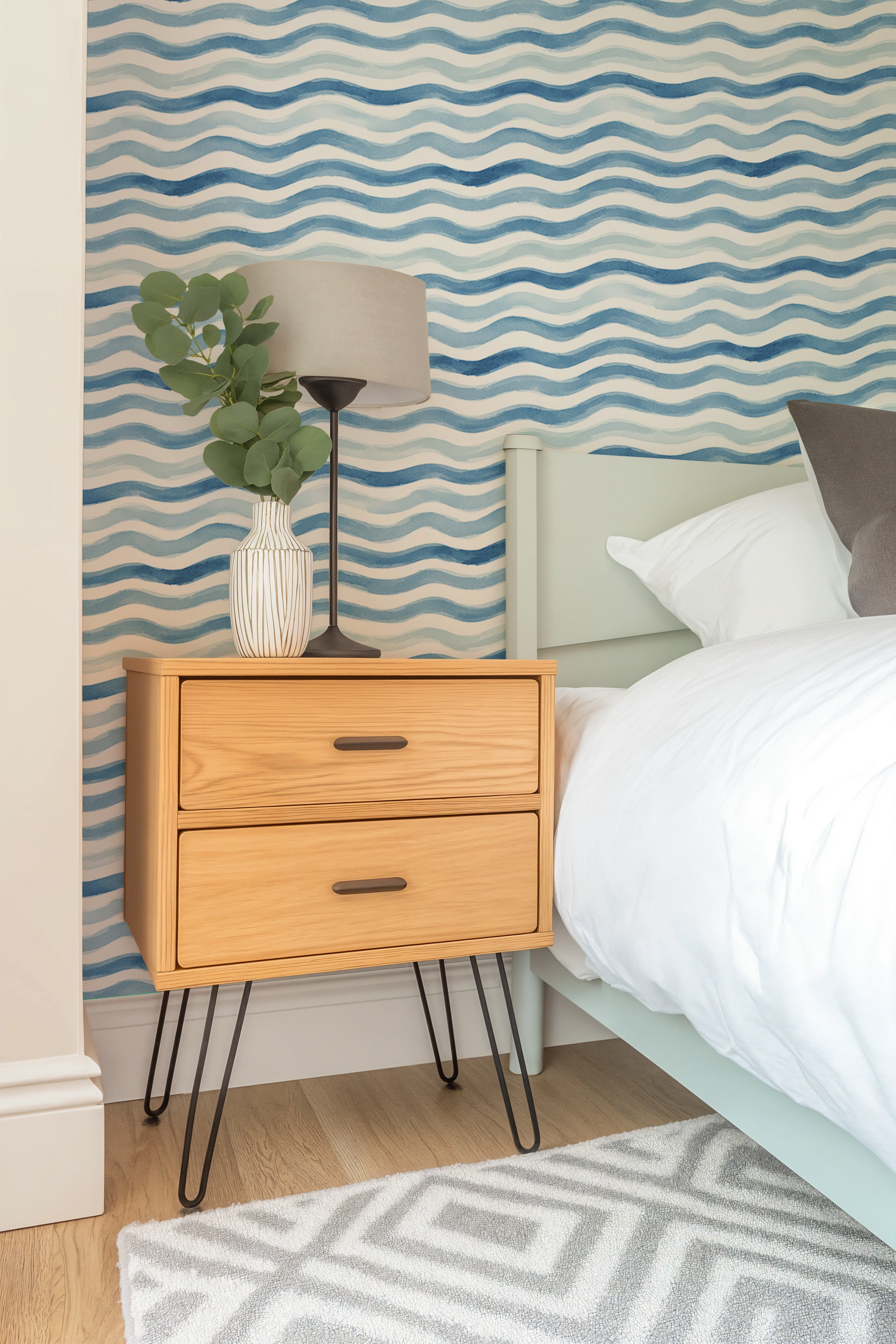 Blue watercolor wave pattern wallpaper in a bedroom with a light wood nightstand, modern lamp, and green plant beside a white