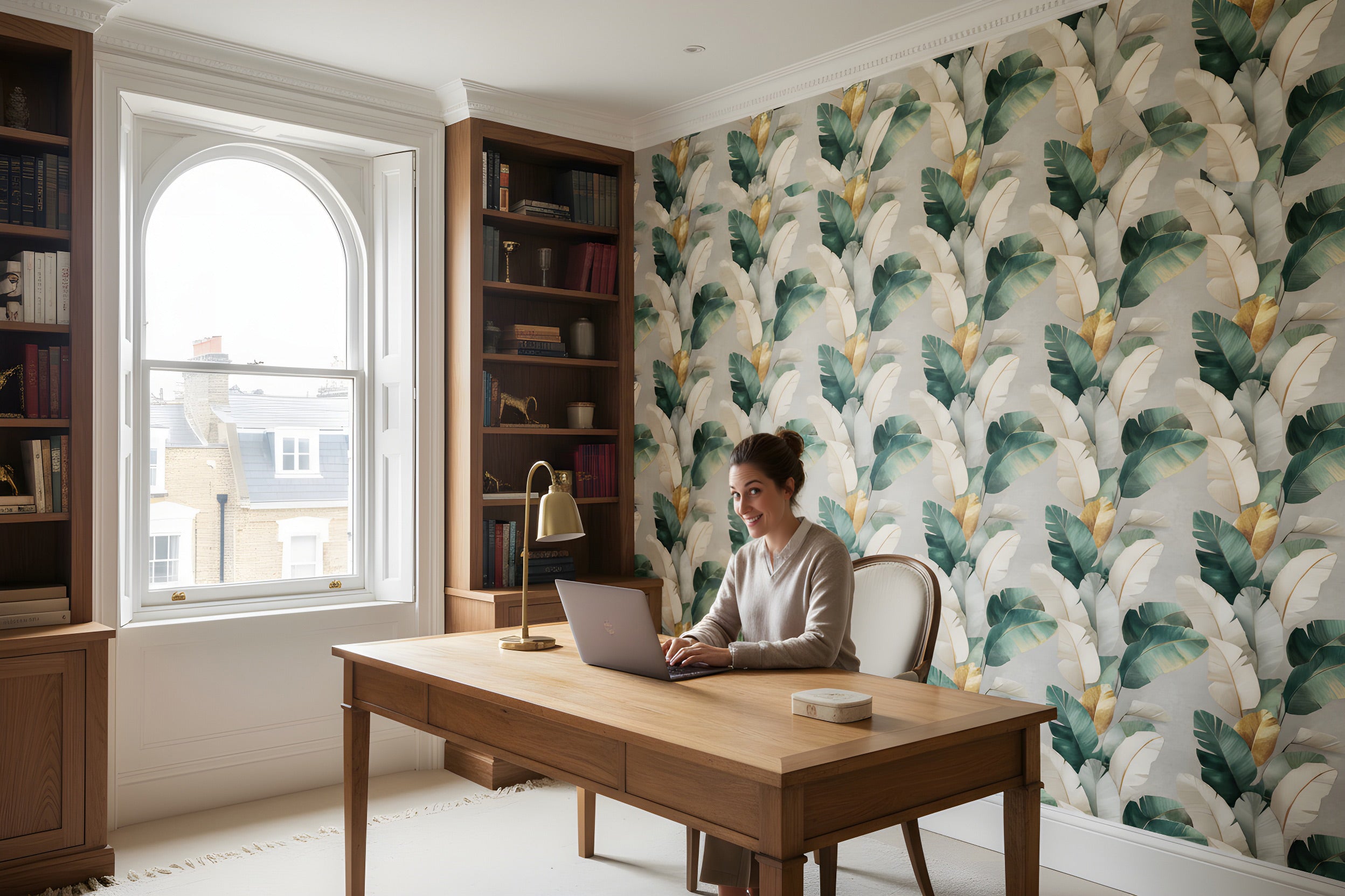 A woman sits at a wooden desk in a bright home office with tropical banana leaf wallpaper in green, teal, cream and gold accents, flanked by tall